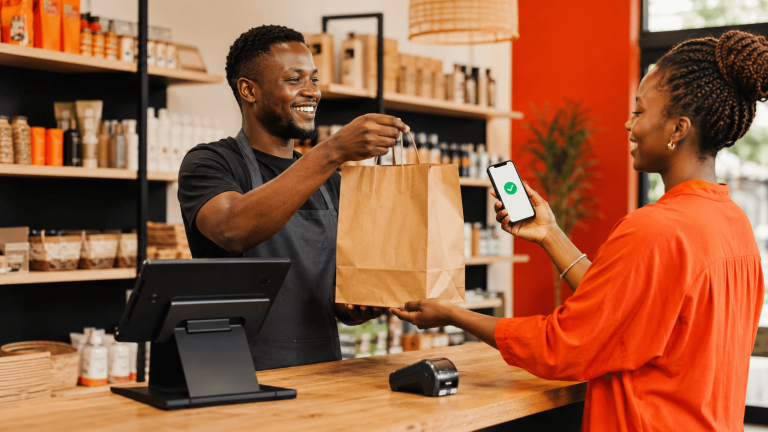 Customer pays with a mobile device while the cashier hands over a brown paper bag at a shop counter.